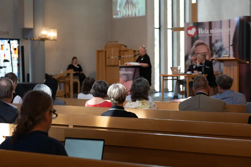 Deltagare på Hela Människans årsstämma i kyrka, med ryggar på personer och talare vid talarstolen i bakgrunden.